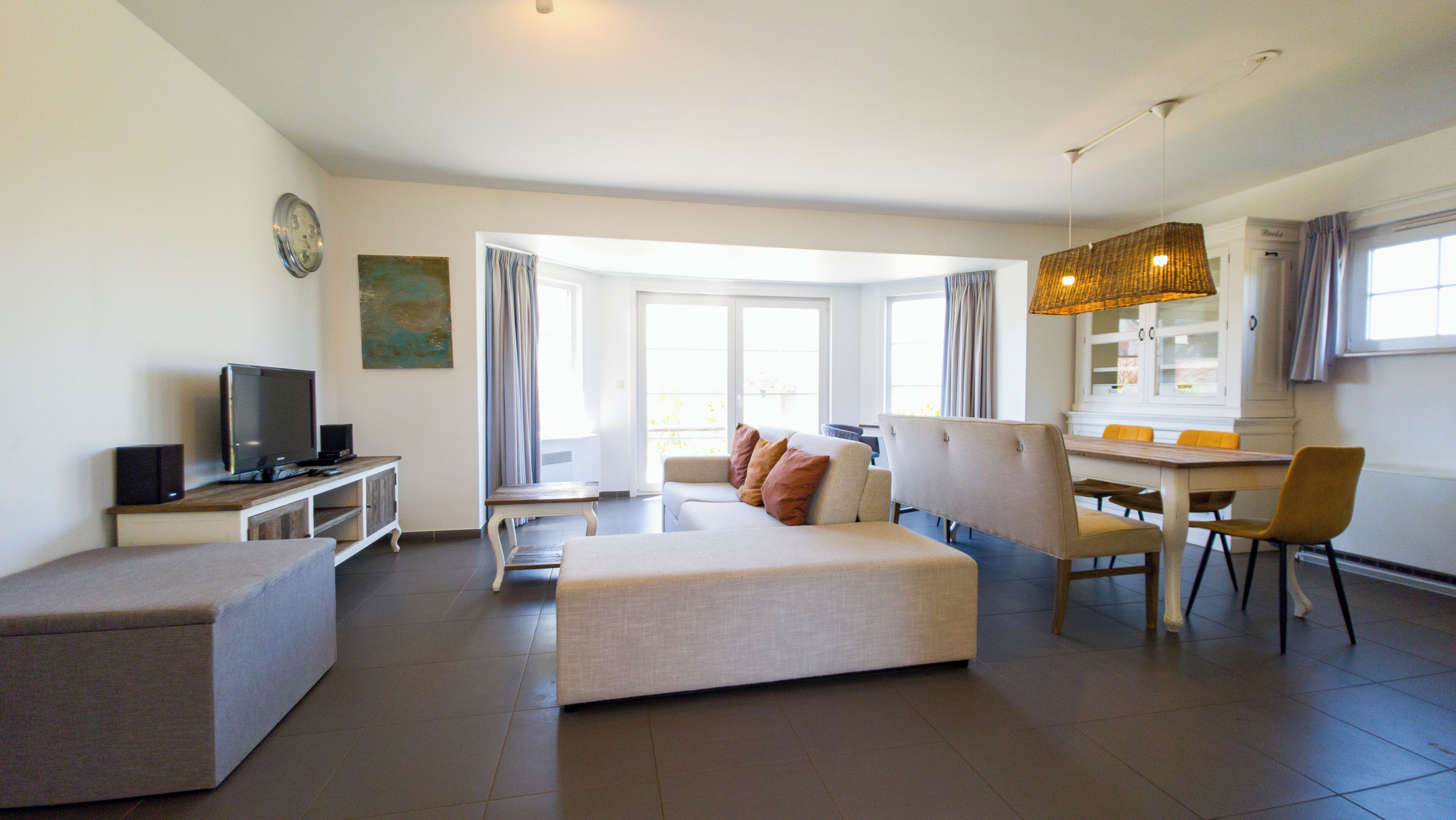 Een moderne woonkamer met een bank, een voetenbankje, een eettafel, stoelen en een televisie.