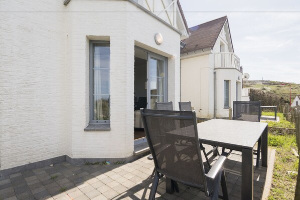 An outdoor patio with a table and chairs, a white building with a window, and a view of the surrounding landscape.