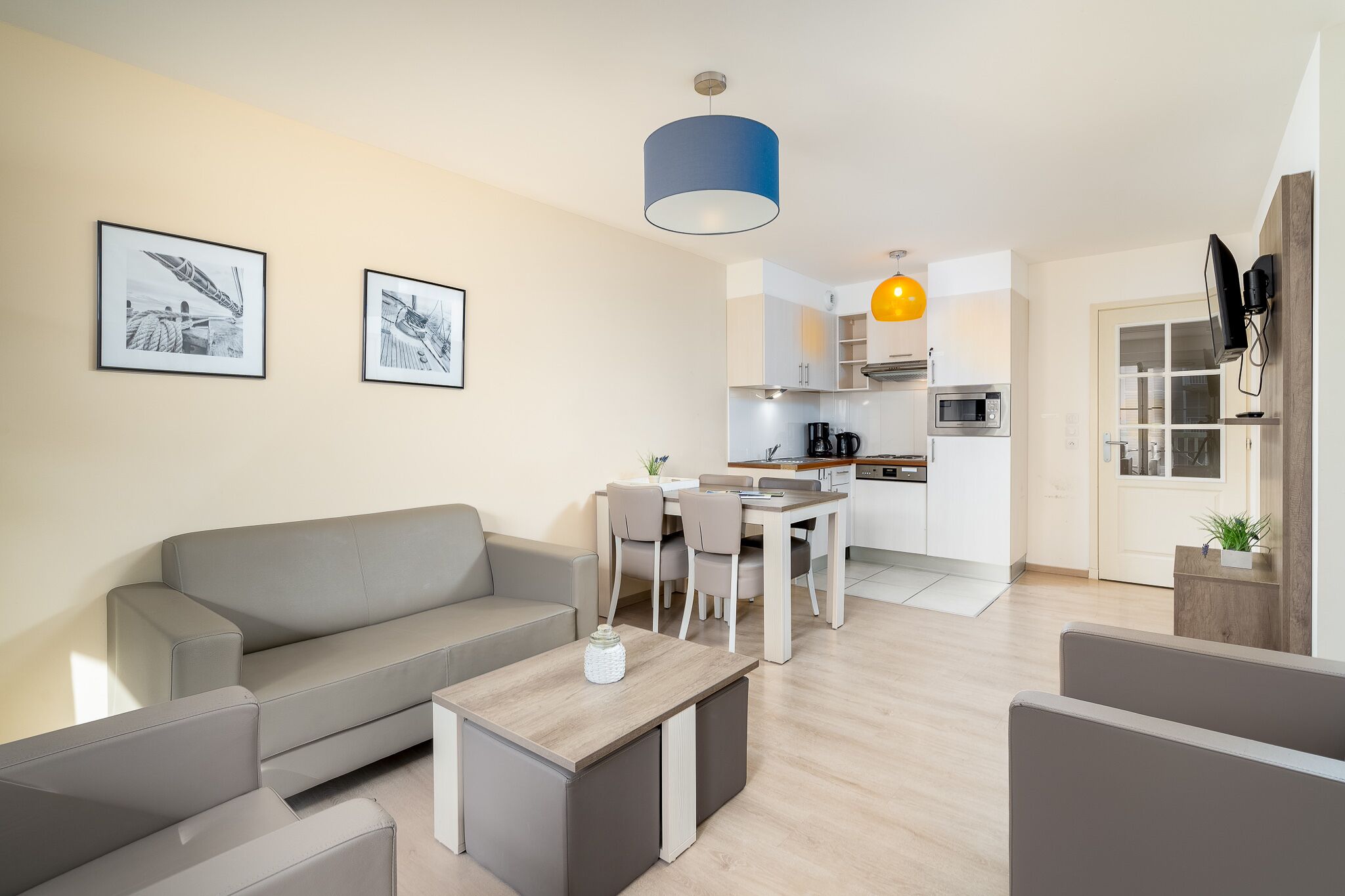 A modern living room with a grey sofa, a coffee table, and a dining area with a table and chairs. There is a kitchen in the background with a microwave and a sink.