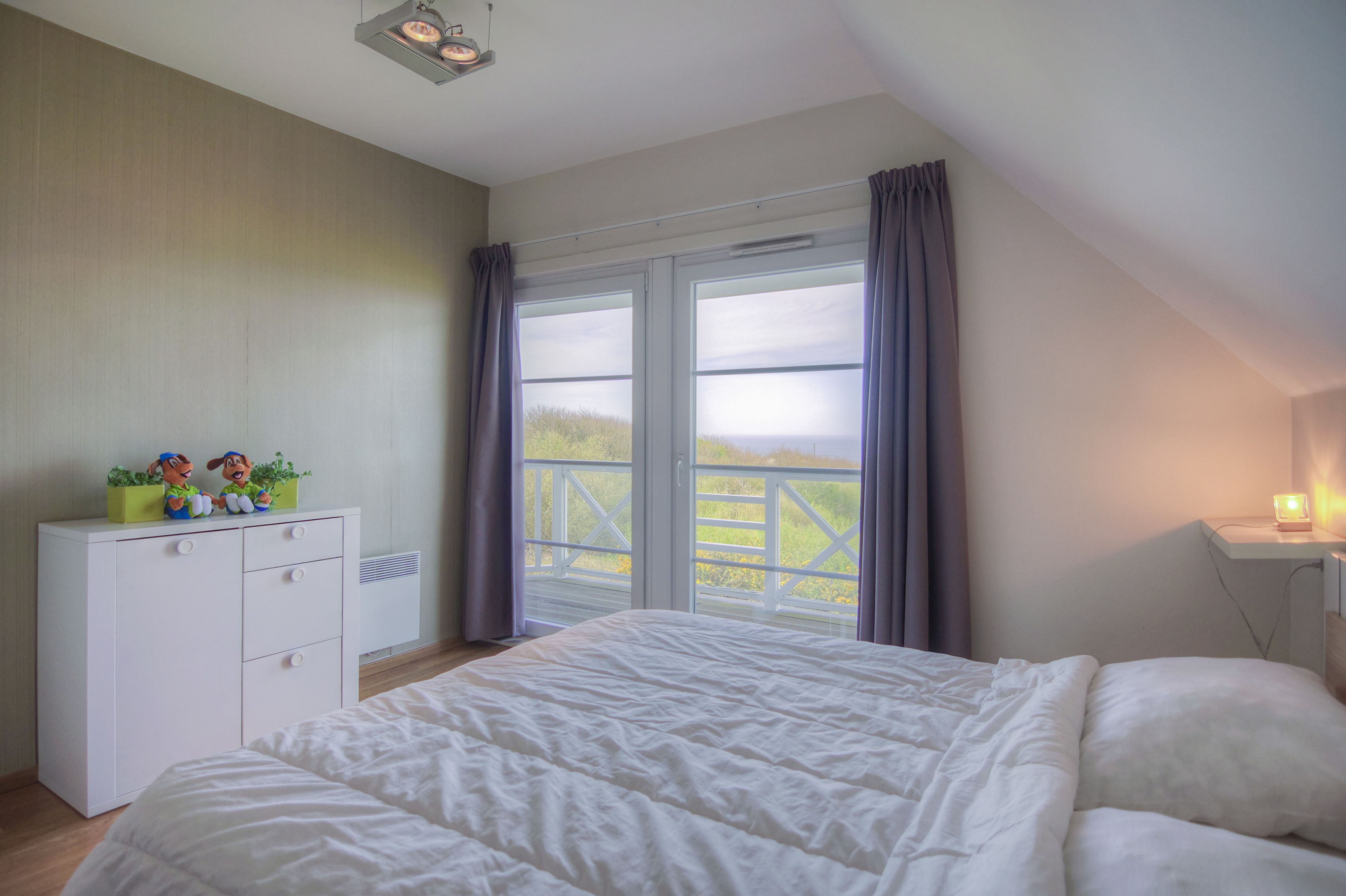 Een slaapkamer met een groot bed, een witte ladekast, een nachtkastje en een raam met gordijnen.