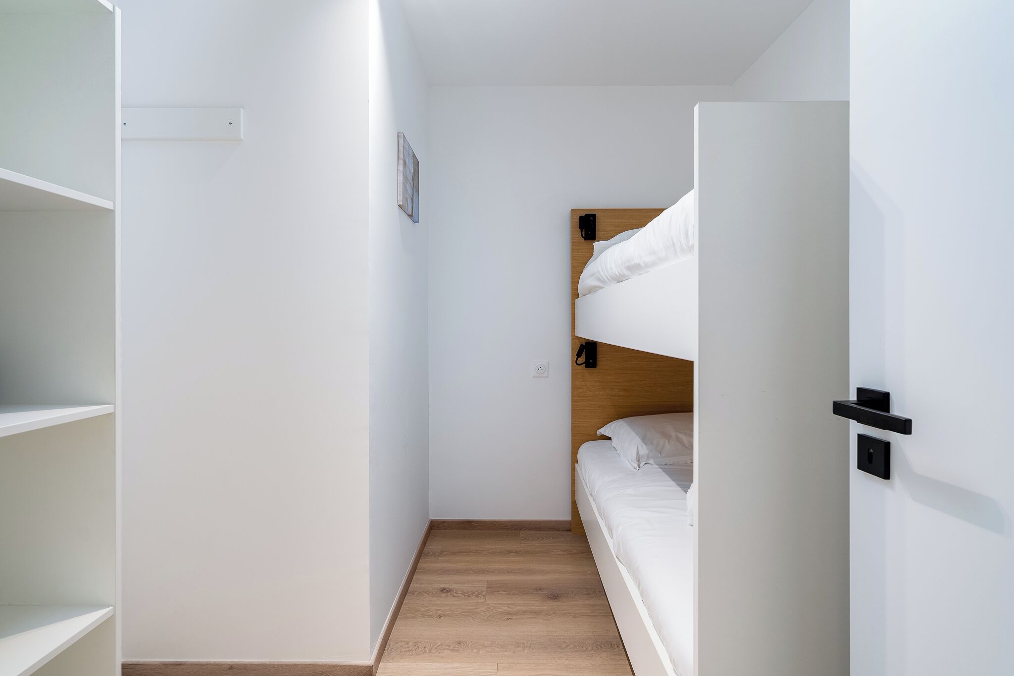 A compact room with a bunk bed, white walls, a wooden floor, and a small shelf unit.