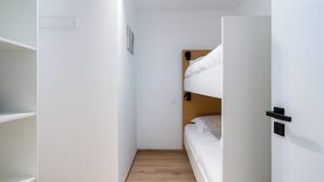 Ein kleines Zimmer mit Etagenbett, weißen Wänden, Holzboden und einer kleinen Regalwand.