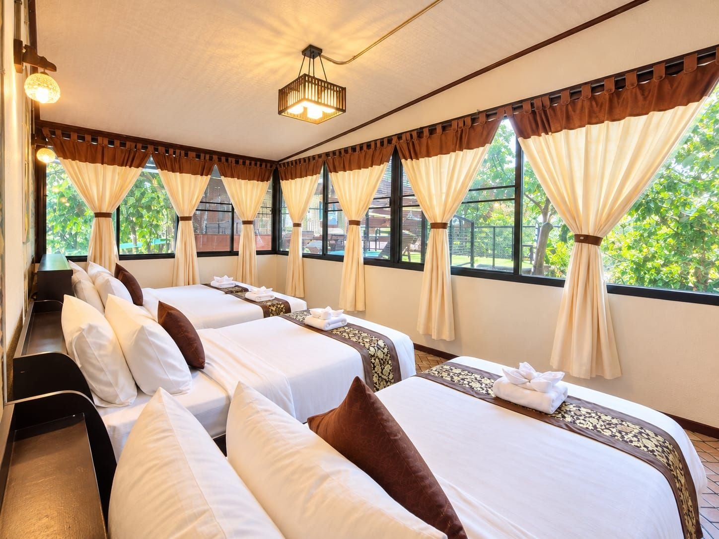 Ein Hotelzimmer mit zwei Betten, großen Fenstern mit Vorhängen, einer Deckenleuchte und Blick auf Grünfläche.