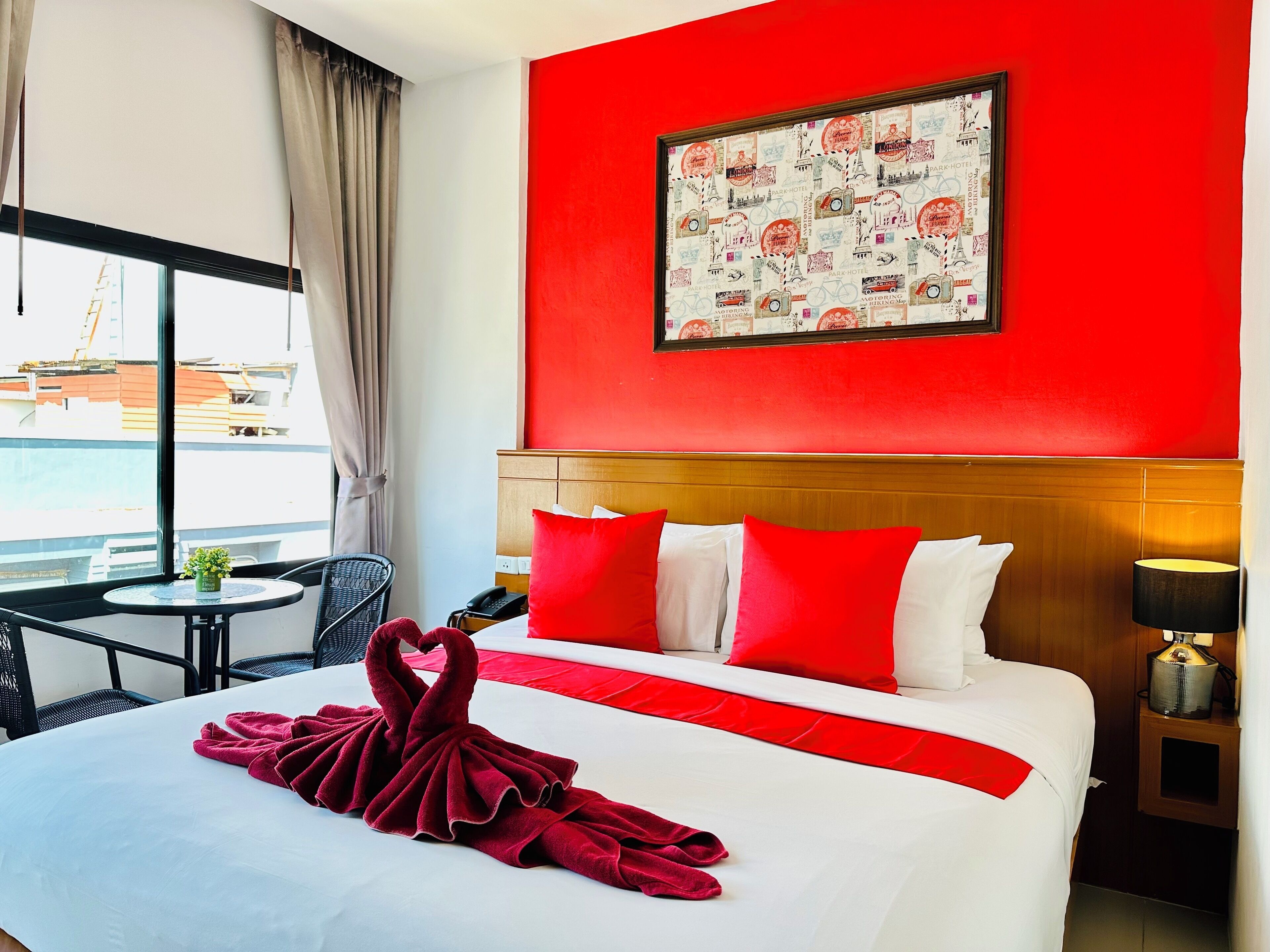 A hotel room with a large bed, red and white pillows, a red and white bedspread, a red towel, a small table with a plant, a chair, a red wall with a framed picture, and a view of a building outside.