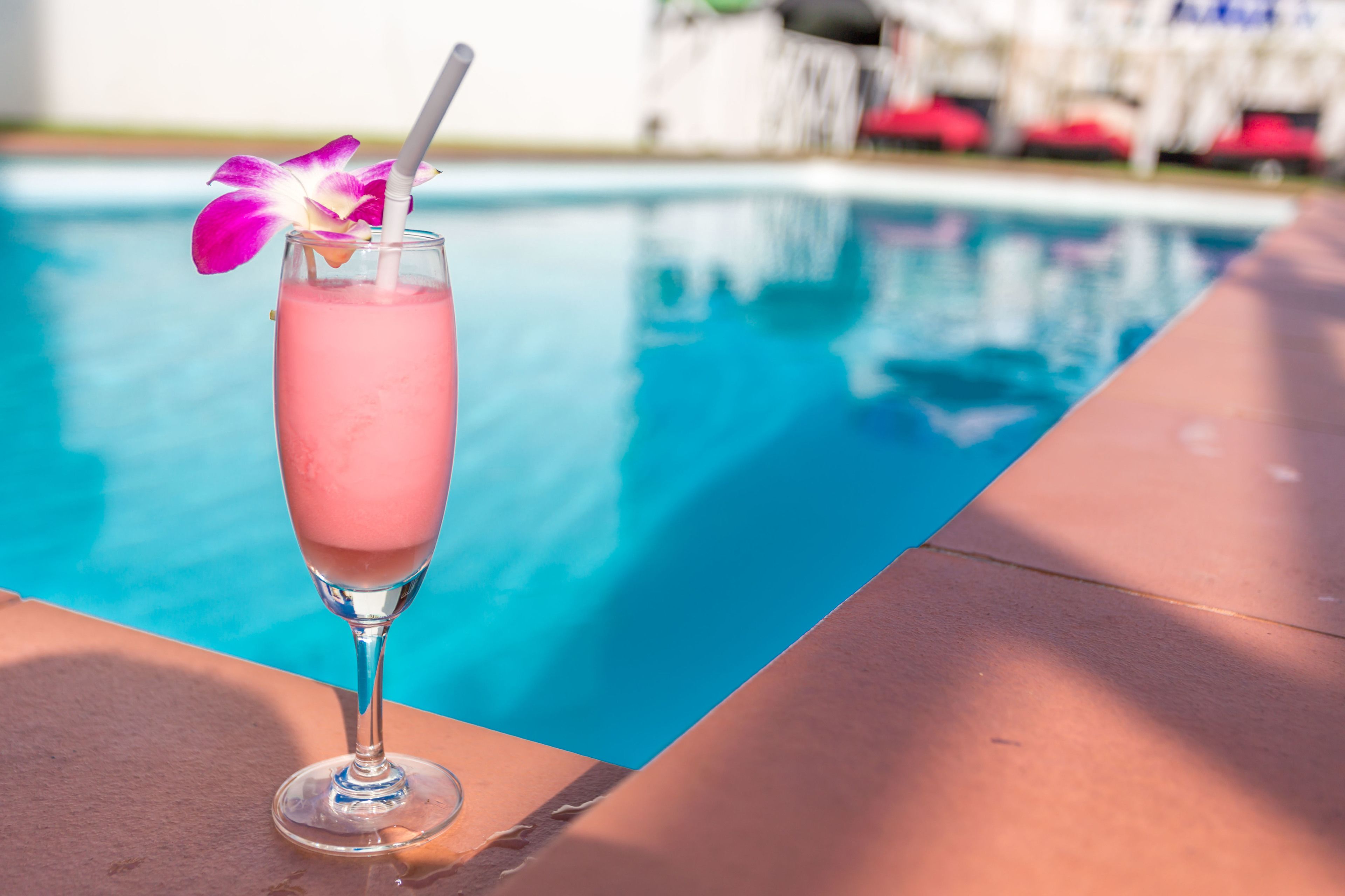 A pink cocktail with a straw and a flower garnish by a poolside.