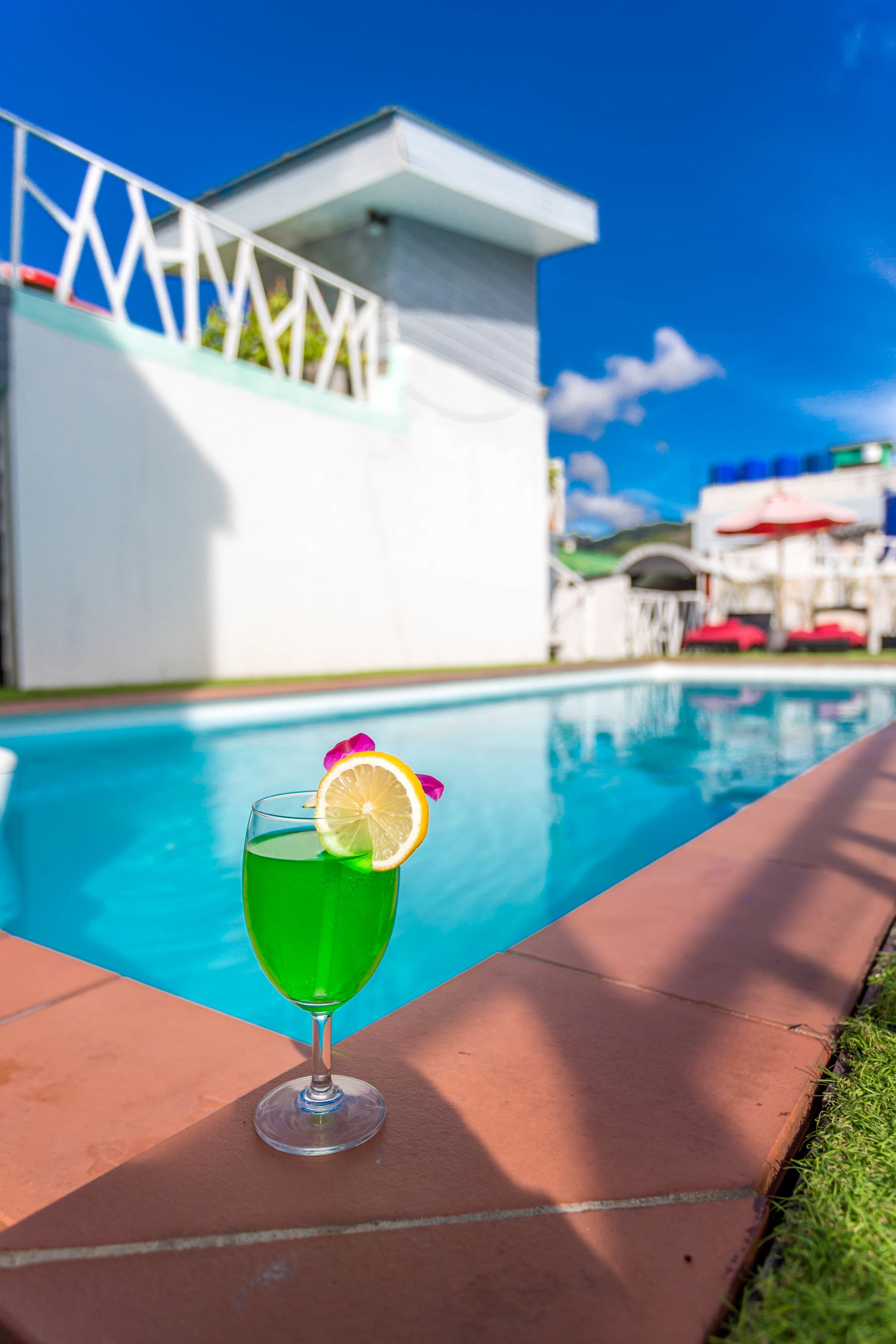 A glass of green drink with a lemon slice and flower on a tiled surface by a pool.