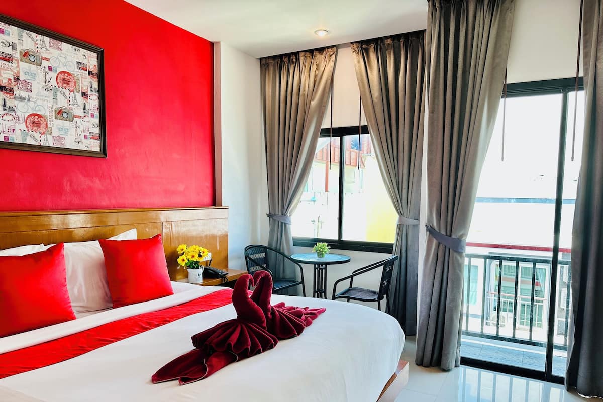 A hotel room with a large bed, red and white bedding, a headboard, a small table with a vase of flowers, a chair, and a balcony with curtains.