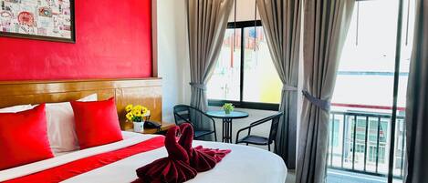 A hotel room with a large bed, red and white bedding, a headboard, a small table with a vase of flowers, a chair, and a balcony with curtains.