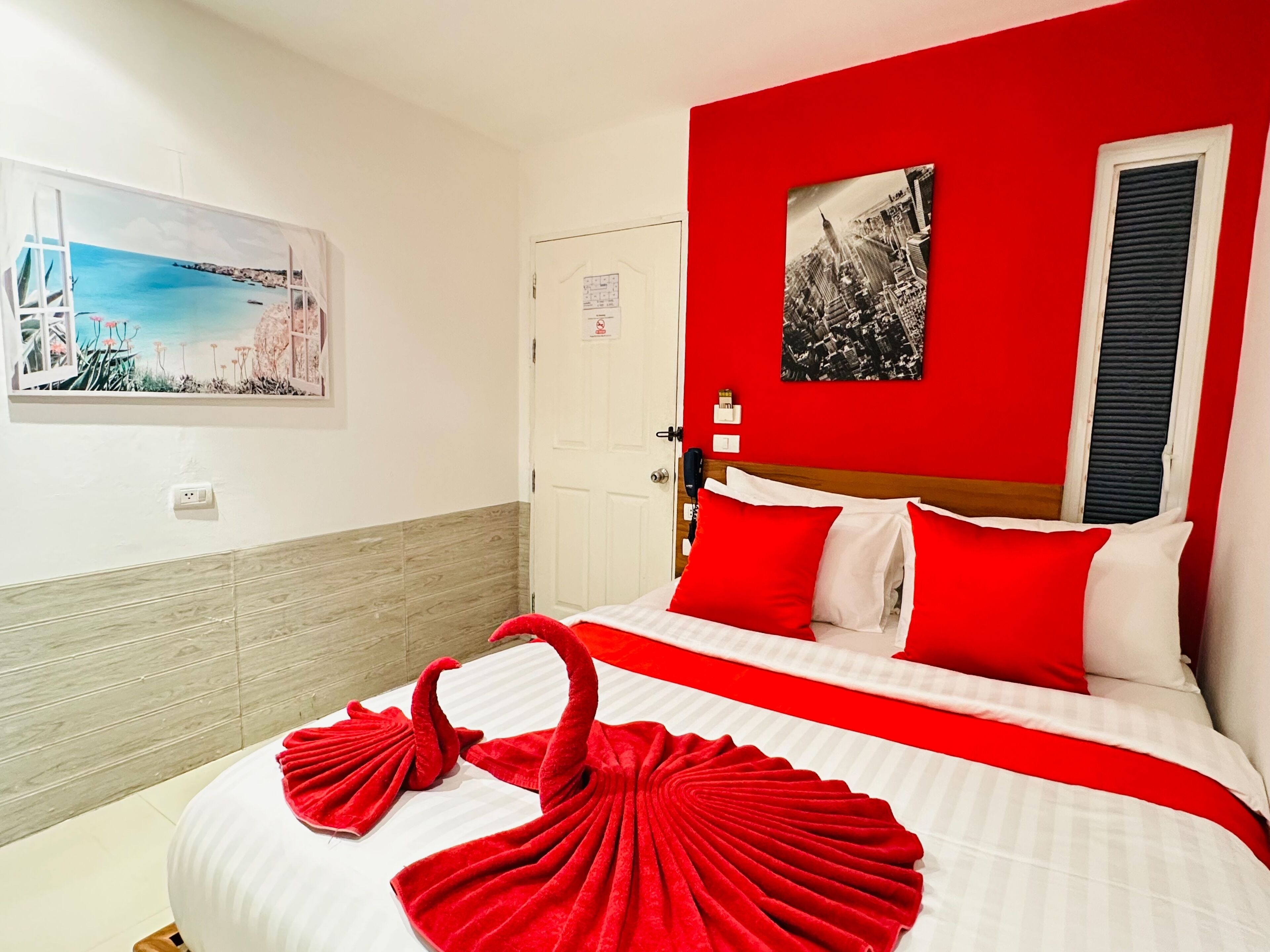 Habitación de hotel con cama, ropa de cama roja y blanca, pared roja y una fotografía de paisaje urbano.