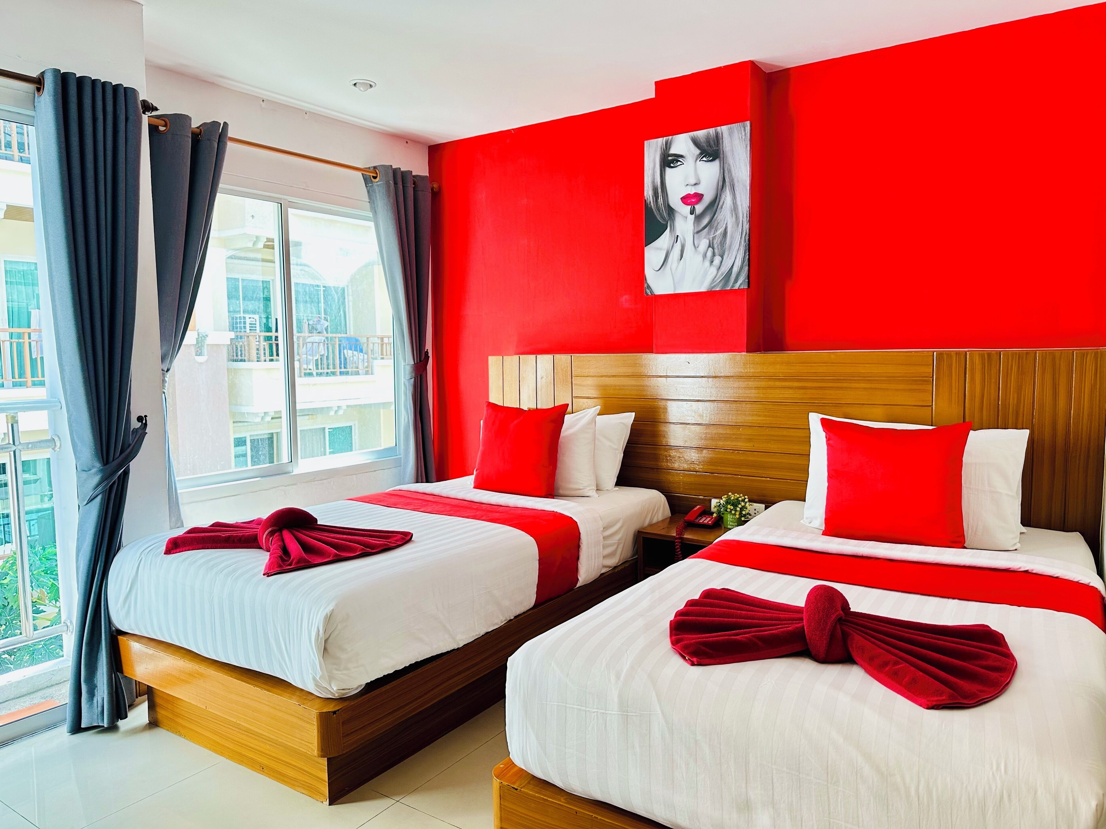 A hotel room with two beds, red and white bedding, a wooden headboard, a red wall, and a window with curtains.