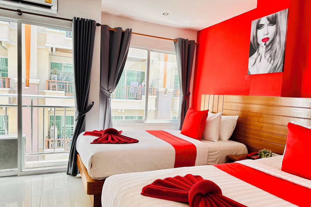 A hotel room with two beds, a red accent wall, and a large window with curtains.