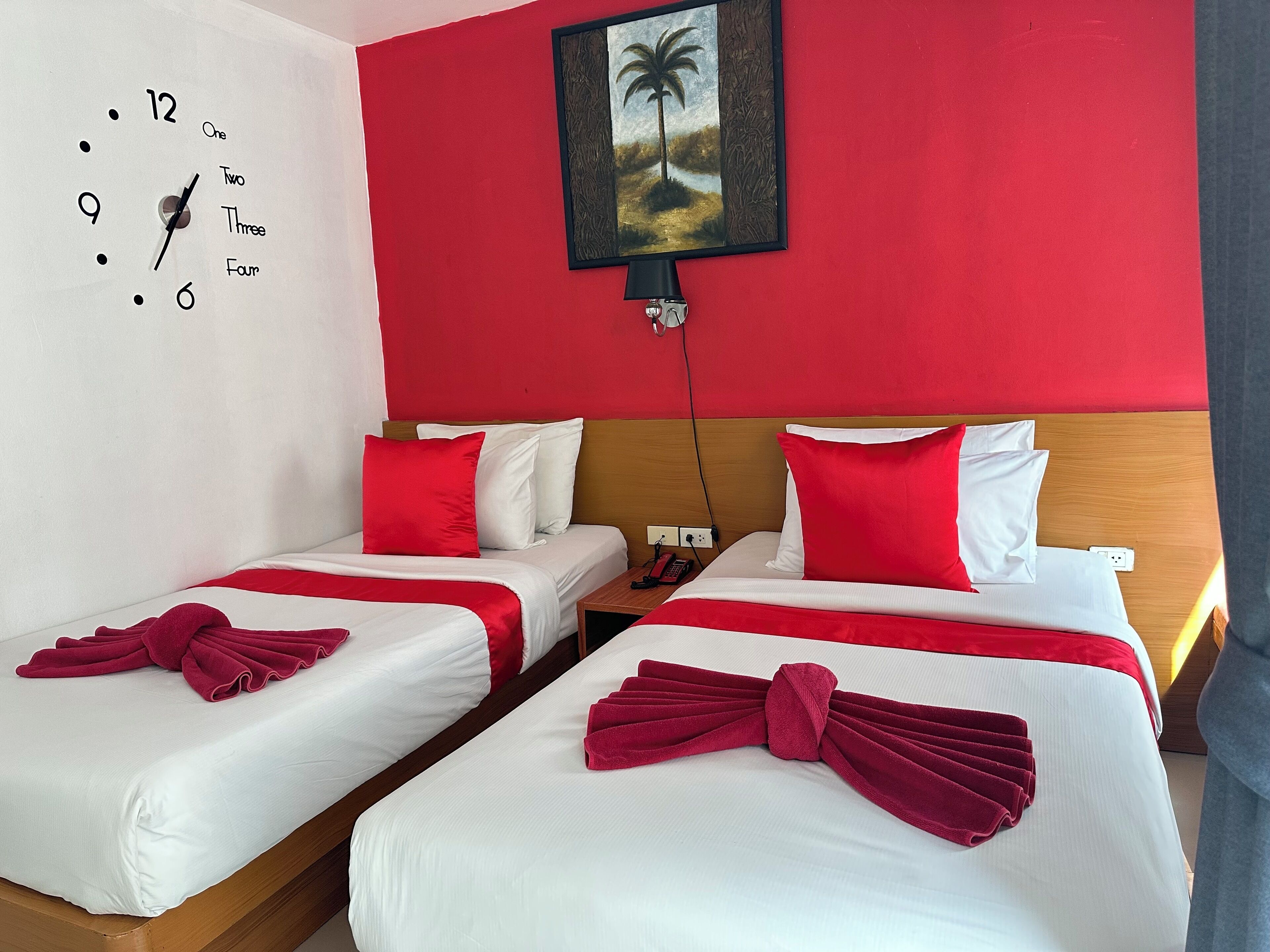 A hotel room with two beds, red and white bedding, a clock on the wall, and a painting of a palm tree.