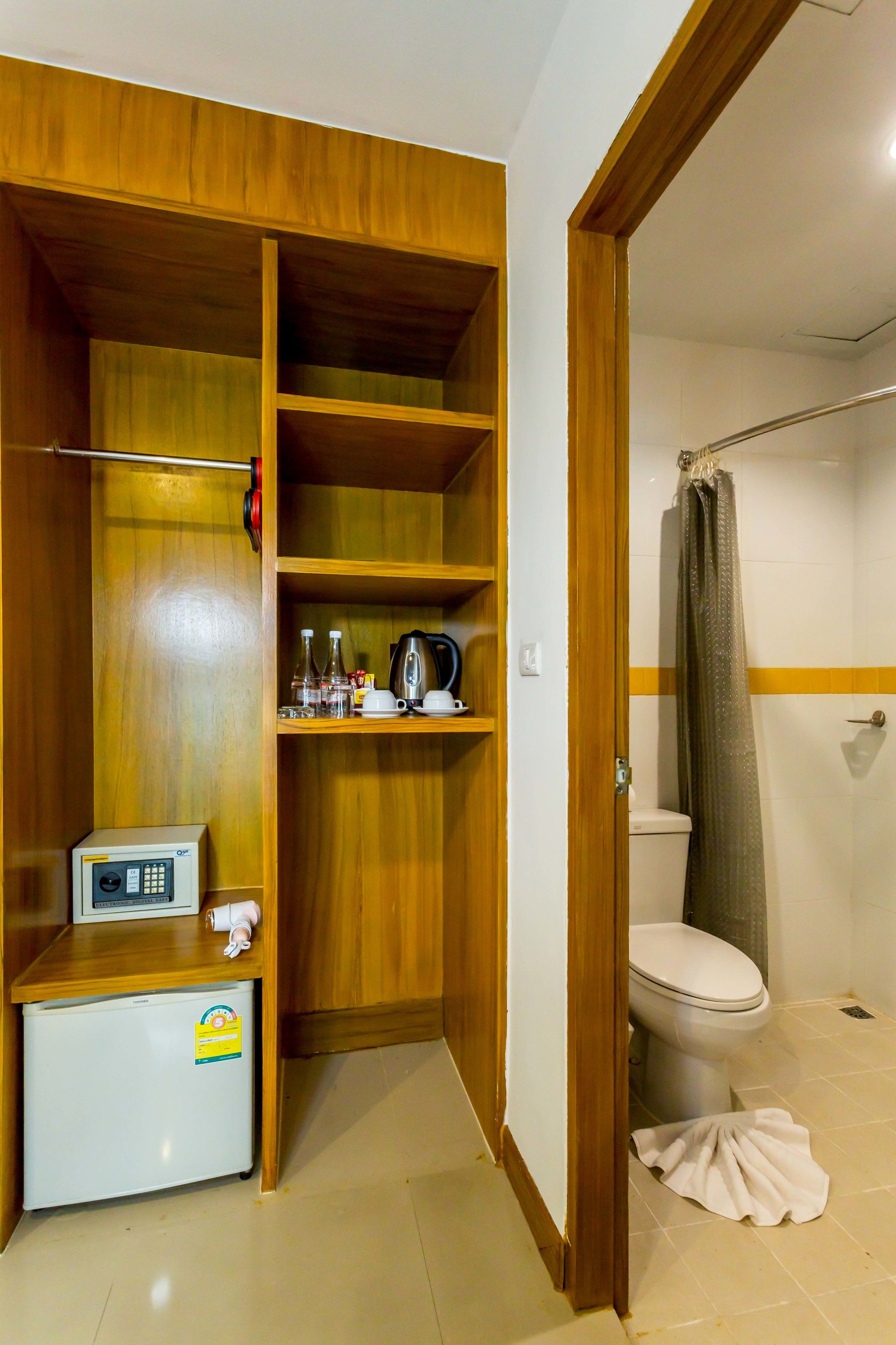 A compact bathroom with a shower, a toilet, a small refrigerator, and a wooden wardrobe with shelves.