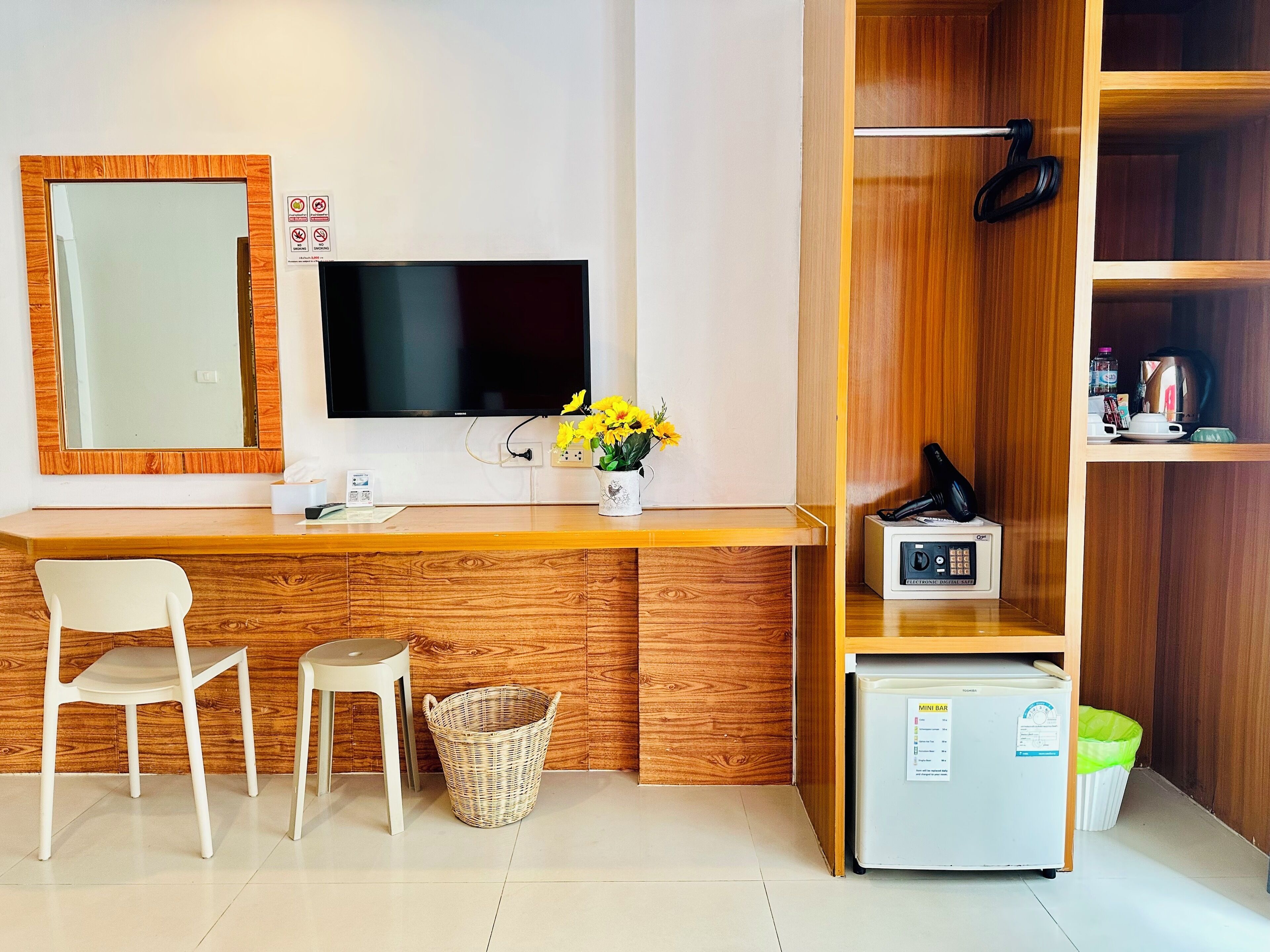A hotel room with a wooden desk, a TV, a chair, a basket, a mini fridge, and a shelf with various items.