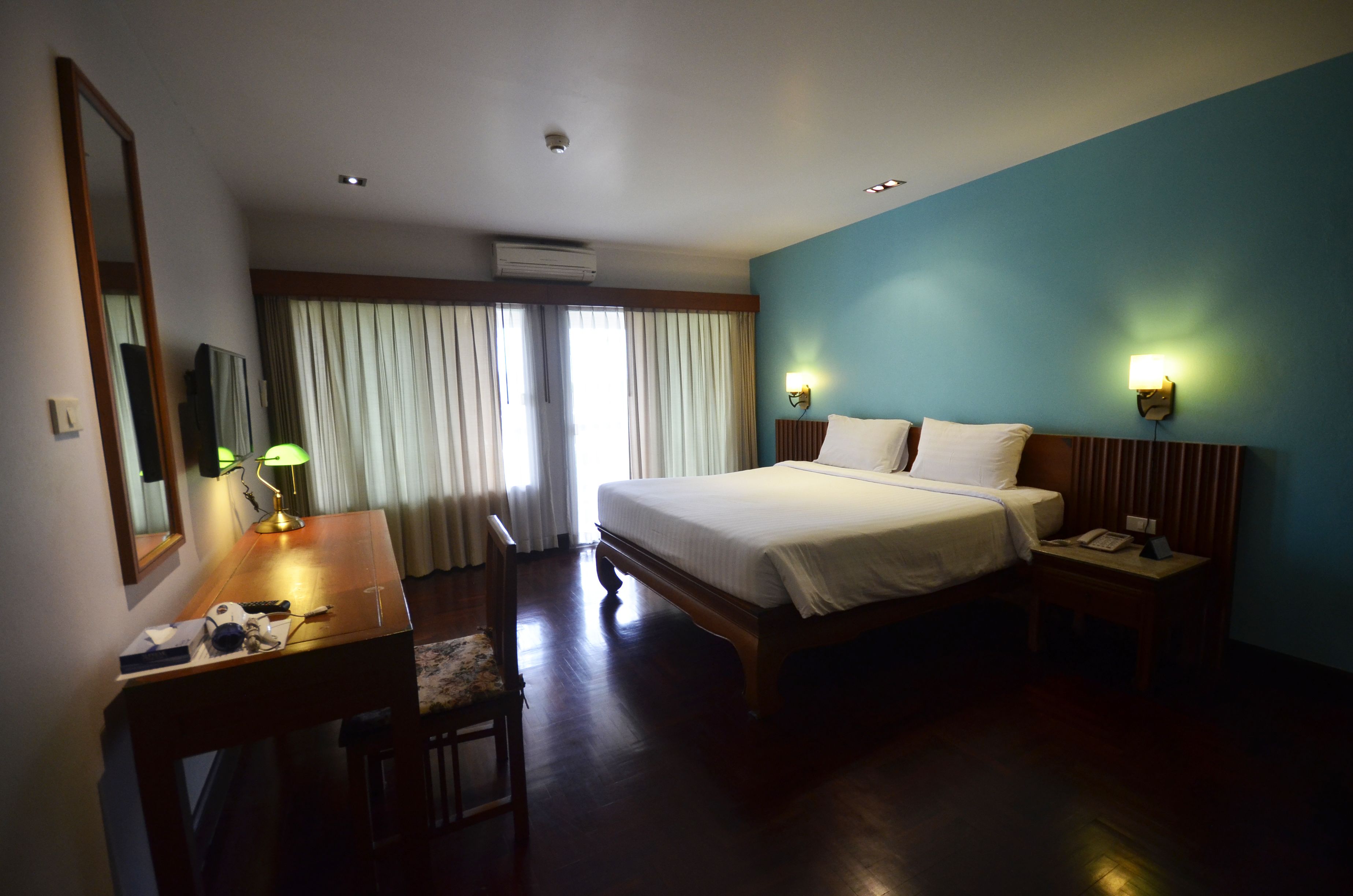 A hotel room with a bed, a desk with a lamp, a chair, a nightstand with a phone, and a wall-mounted light.