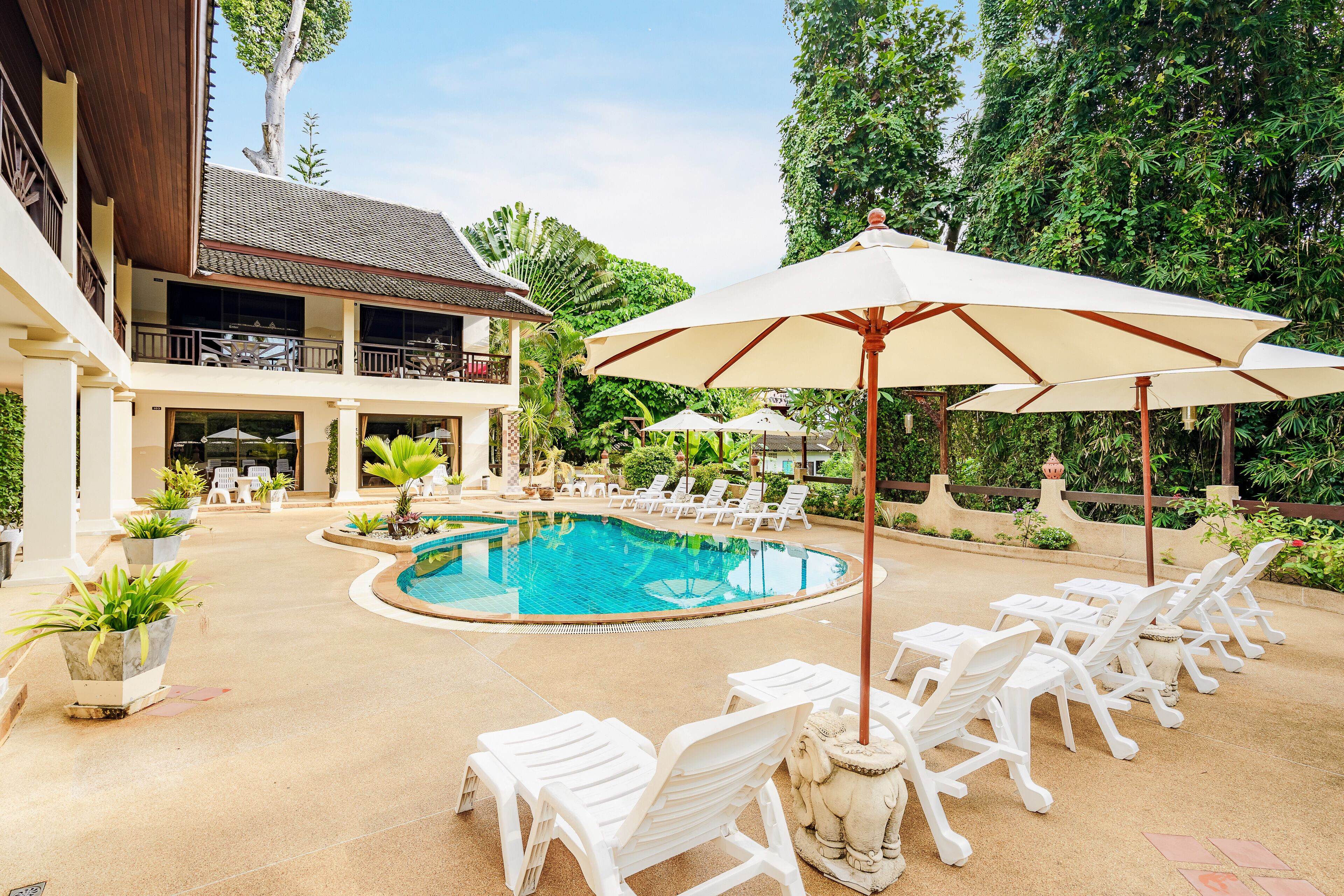 A pool area with lounge chairs and umbrellas, surrounded by lush greenery and a building with a balcony.