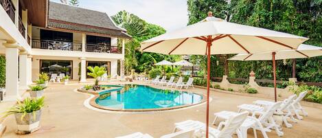 A pool area with lounge chairs and umbrellas, surrounded by lush greenery and a building with a balcony.