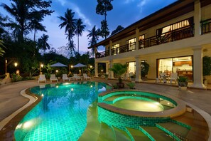 Une piscine au design incurvé, entourée de chaises longues et de parasols, avec un bâtiment doté d’un balcon et d’une cuisine visible à l’arrière-plan.