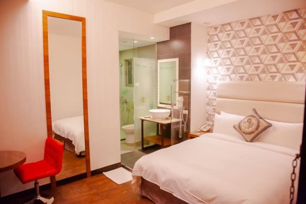 Habitación de hotel con cama, una silla roja, un espejo grande, baño con inodoro y lavamanos, y pared con diseño.