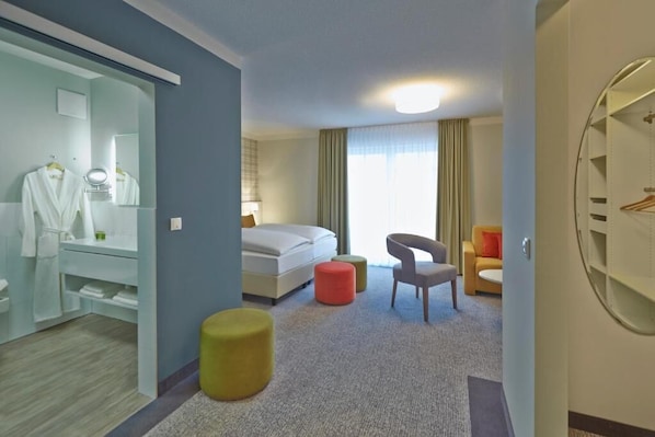 A hotel room with a bed, a chair, a small ottoman, a mirror, a wardrobe, and a circular mirror on the wall.