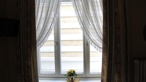 A room with a window featuring white curtains, a small table with a vase of yellow flowers, and a view of a building outside. - Hotel-Pension Fink (Hamburg)