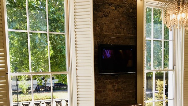 A hotel room with large windows, a flat-screen TV mounted on a brick wall, and a bed with white linens. - The Melville Hotel by Rokxy - Kings Cross (London)