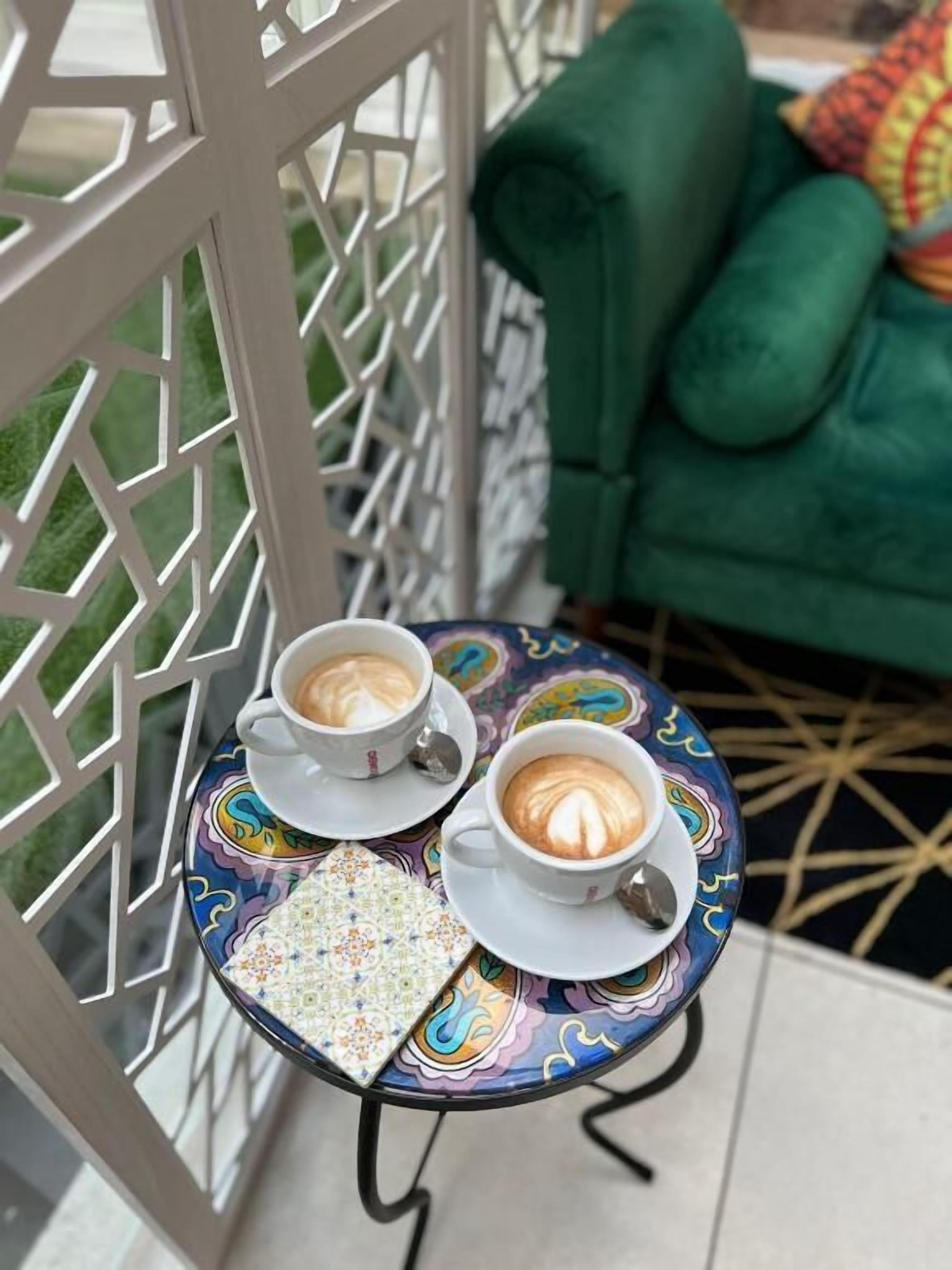 Uma mesa redonda pequena com duas chávenas de café brancas, um tabuleiro estampado e um sofá verde ao fundo.