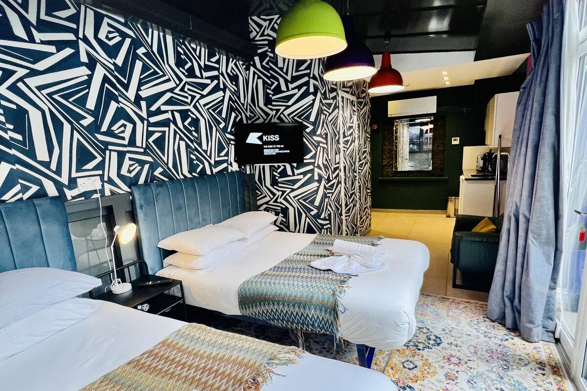 A modern hotel room with a patterned wall, a massage table, a bed, and a colorful rug.