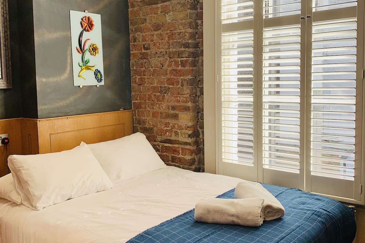 A neatly made bed with white linens and a blue checkered blanket, a brick wall, and a window with blinds.
