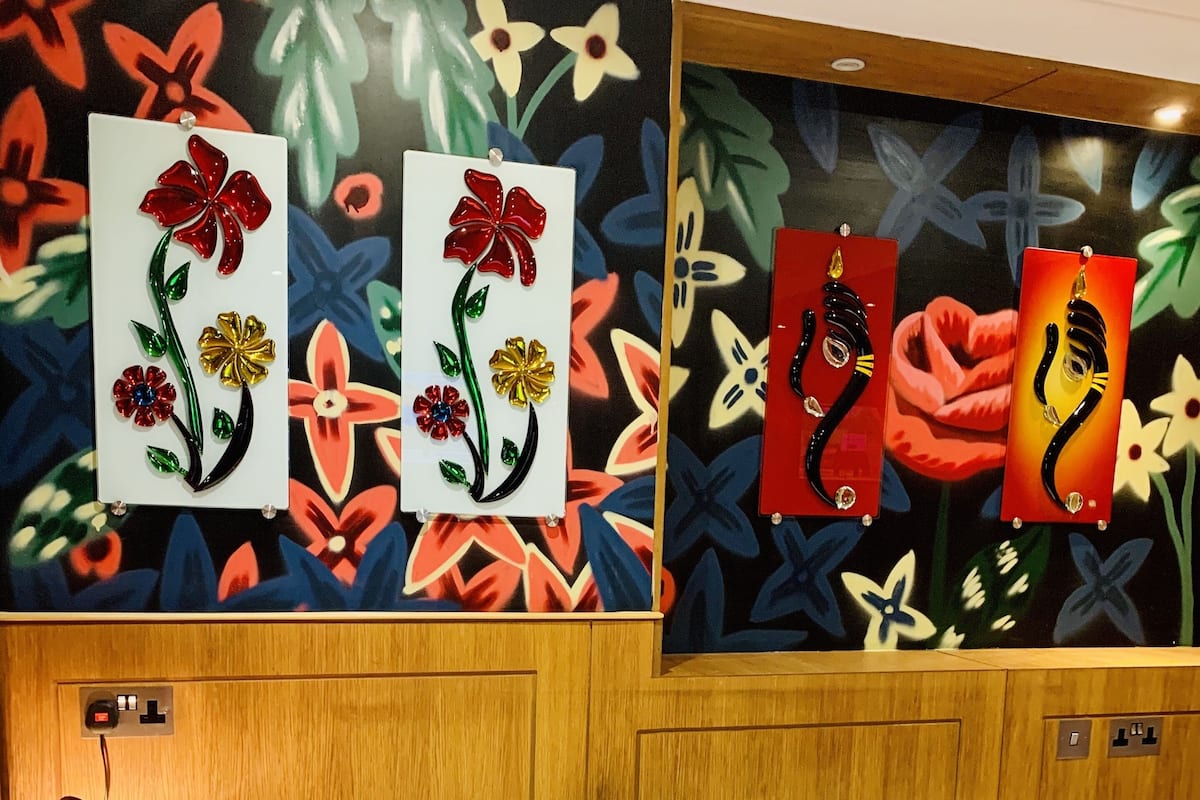 A hotel room with a bed, a nightstand with a lamp, and wall art featuring floral designs.
