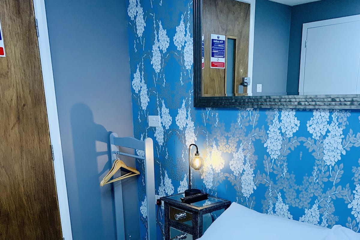 A hotel room with a bed, a nightstand with a lamp, a mirror, and a wall with a floral pattern.