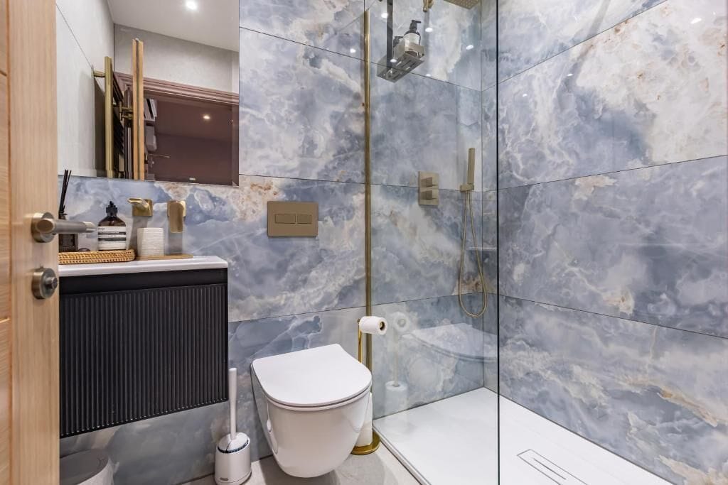 A modern bathroom with a marble wall, a toilet, a shower, and a sink with a gold faucet.