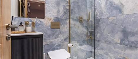 A modern bathroom with a marble wall, a toilet, a shower, and a sink with a gold faucet.