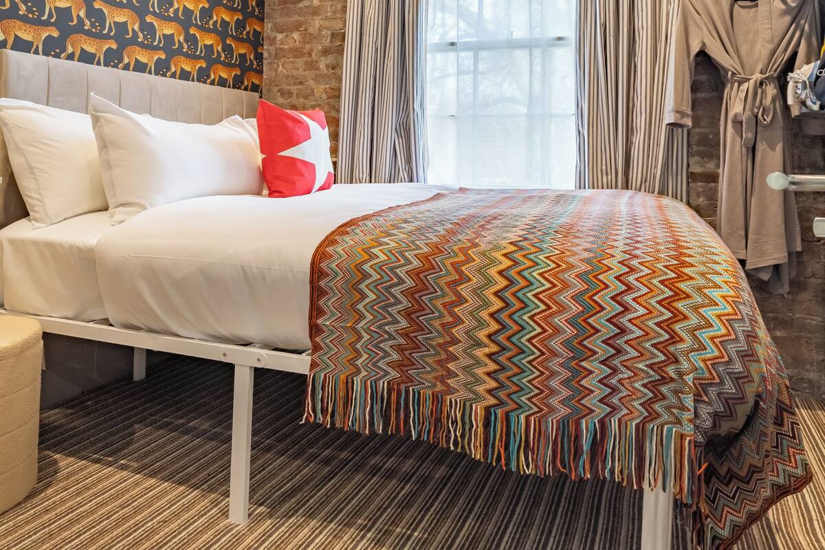 Quarto de hotel com cama, parede estampada, janela com cortinas, cabideiro e detector de incêndio no teto.
