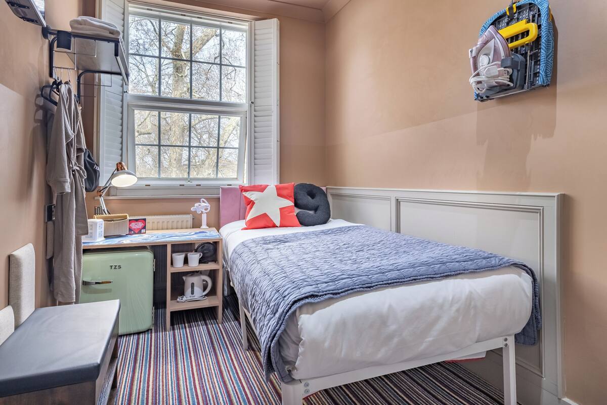 Quarto pequeno com cama, escrivaninha, radiador e janela.