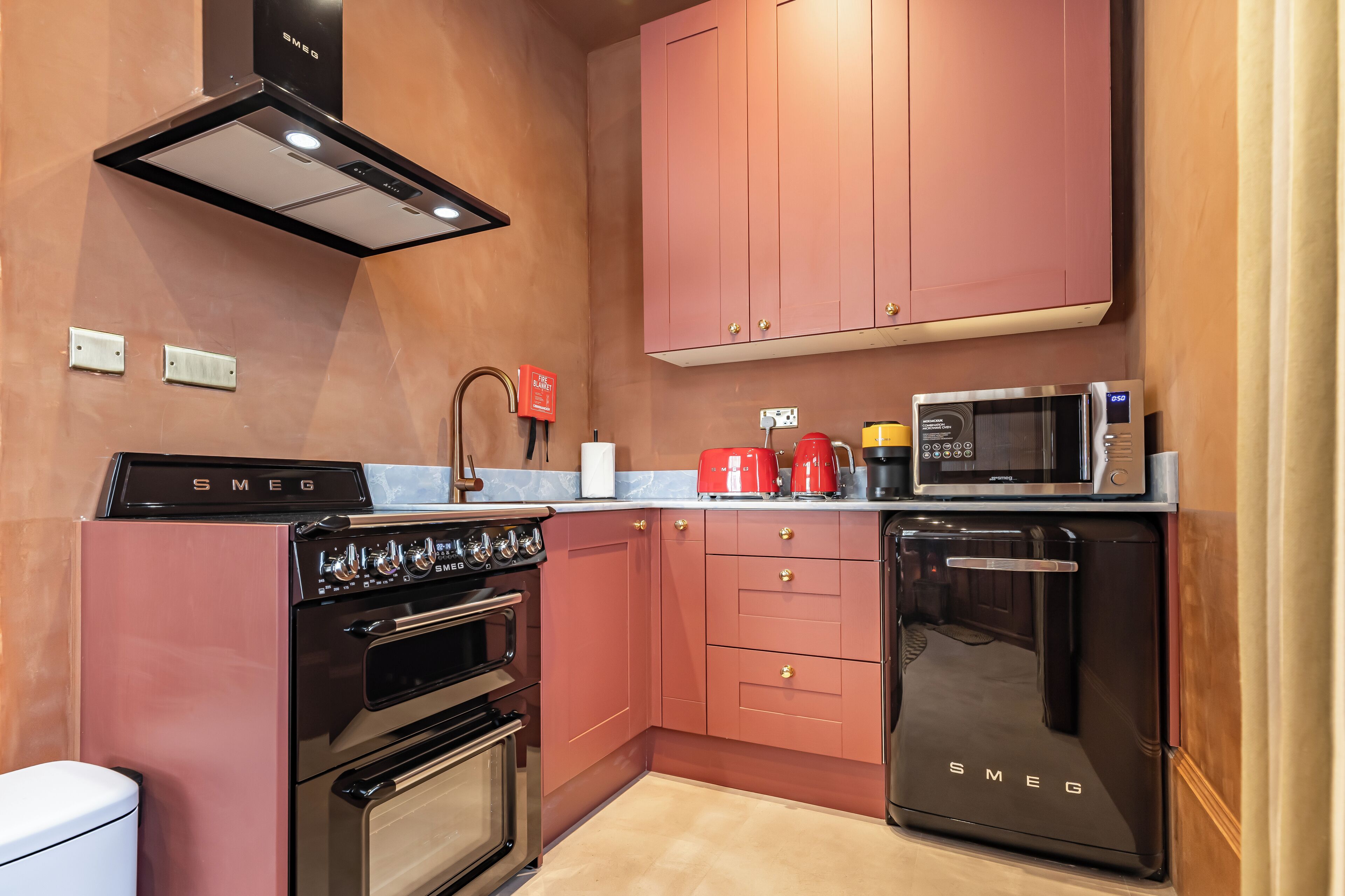 Una cocina con armarios rosa, una campana extractora negra, microondas, cafetera y lavavajillas.