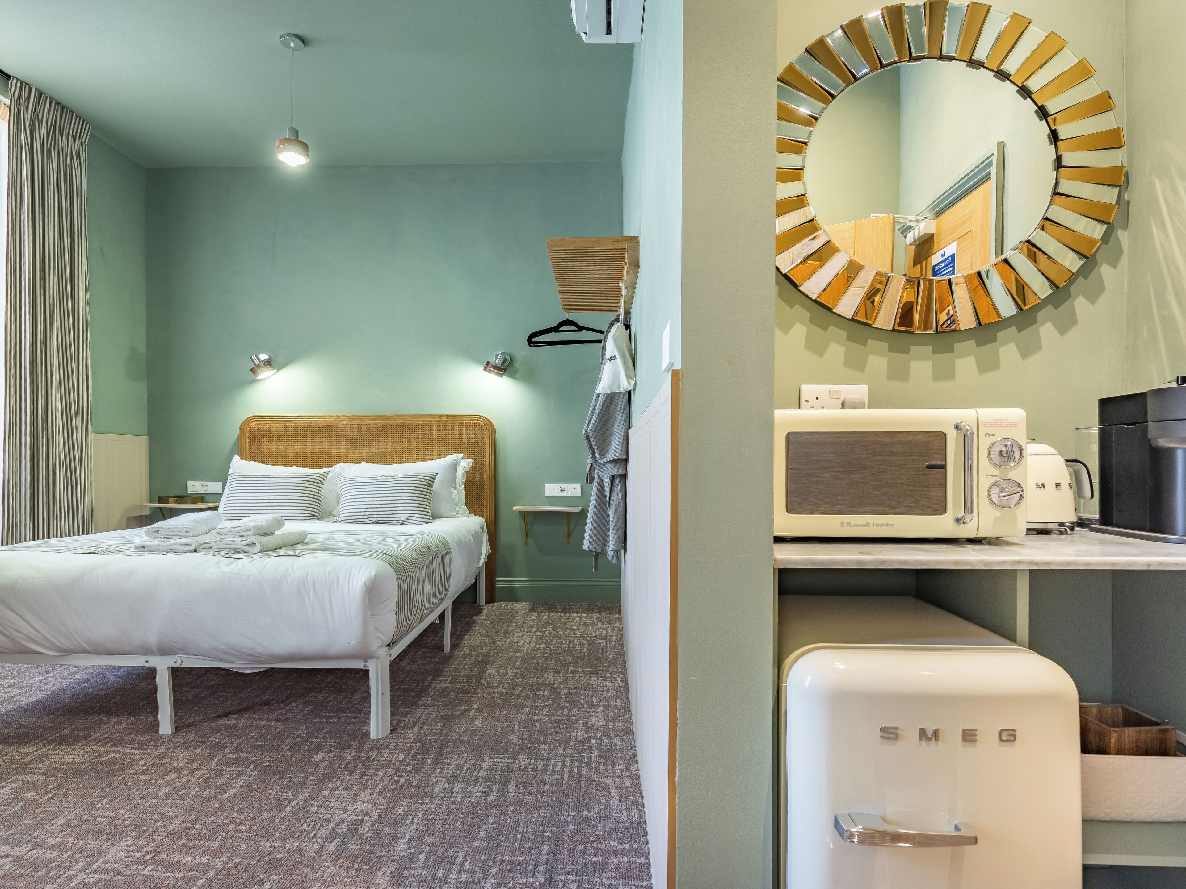 A hotel room with a bed, a bedside table, a mirror, a wall-mounted heater, a towel rack, and a kitchenette with a microwave and toaster.