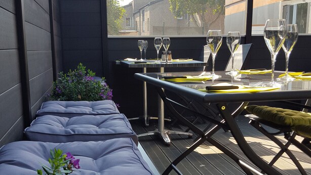 A patio with cushioned seating, a table set for dining, and a backdrop of a building and trees. - Fasthôtel Dunkerque (Grande-Synthe)
