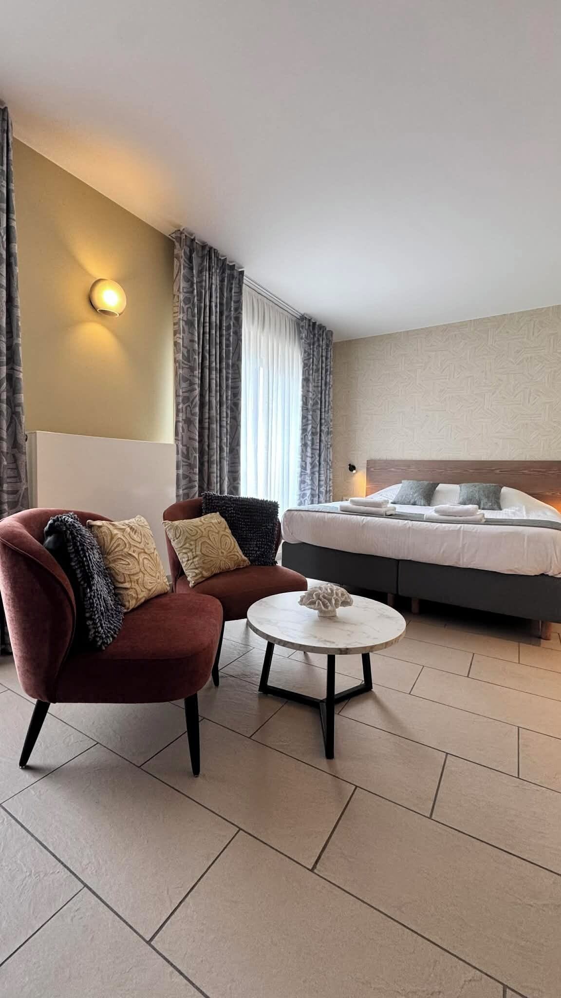 Une chambre d’hôtel avec un lit, un fauteuil rouge, une petite table ronde et une lampe fixée au mur.