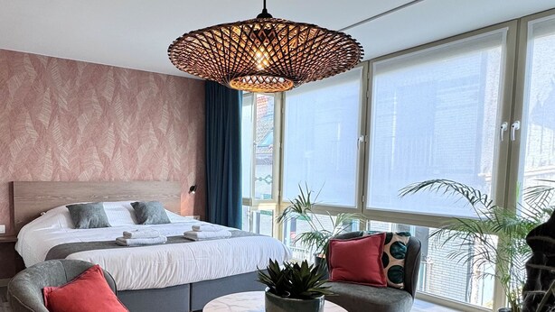 A modern bedroom with a bed, two armchairs, a coffee table, a hanging light fixture, and large windows. - Hotel José (Blankenberge)