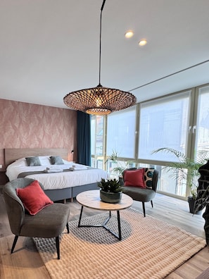 A modern bedroom with a bed, two armchairs, a coffee table, a hanging light fixture, and large windows. - Hotel José (Blankenberge)