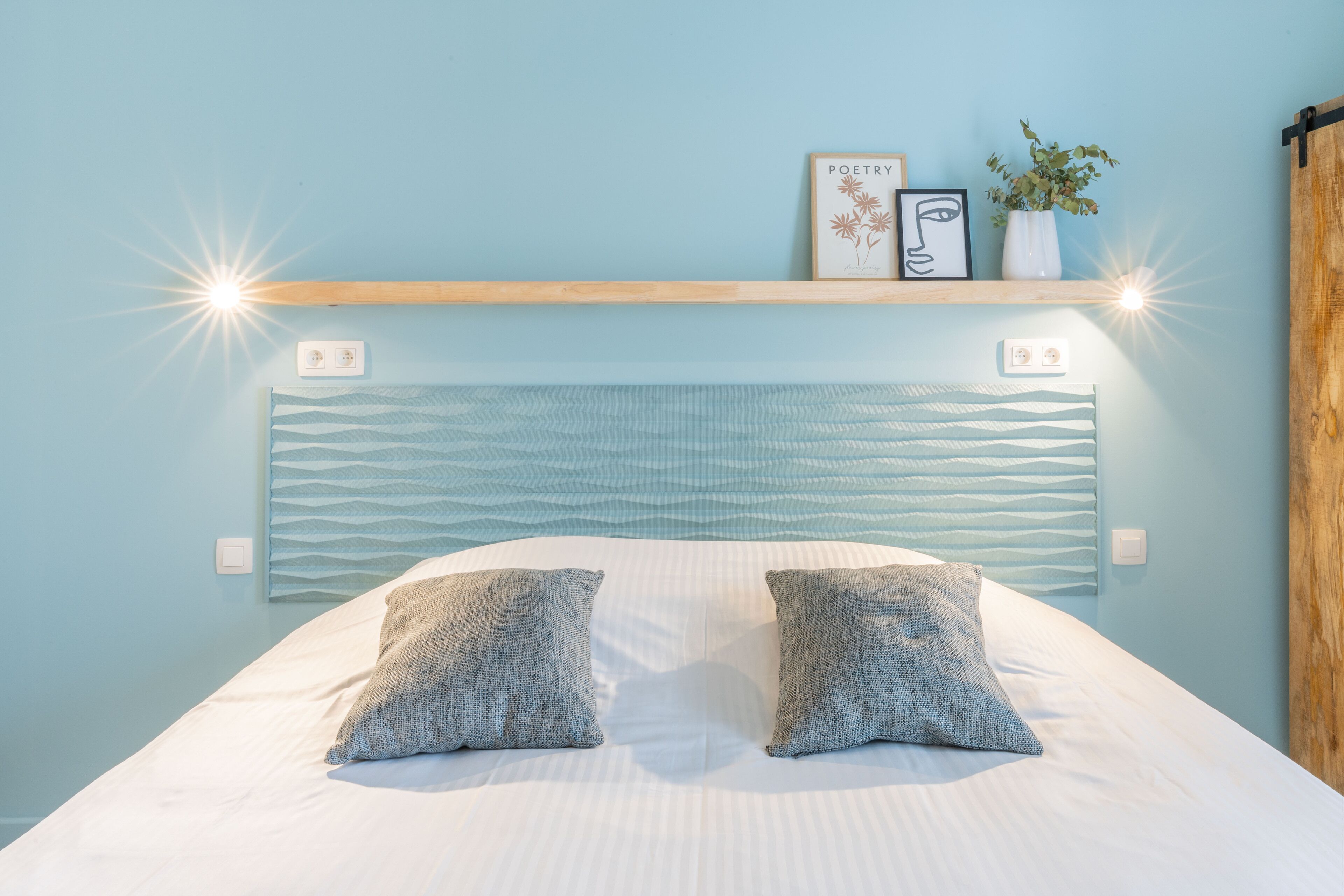 Une chambre à coucher avec un lit, deux oreillers, une tête de lit, une étagère avec un tableau encadré, une petite plante et un luminaire.