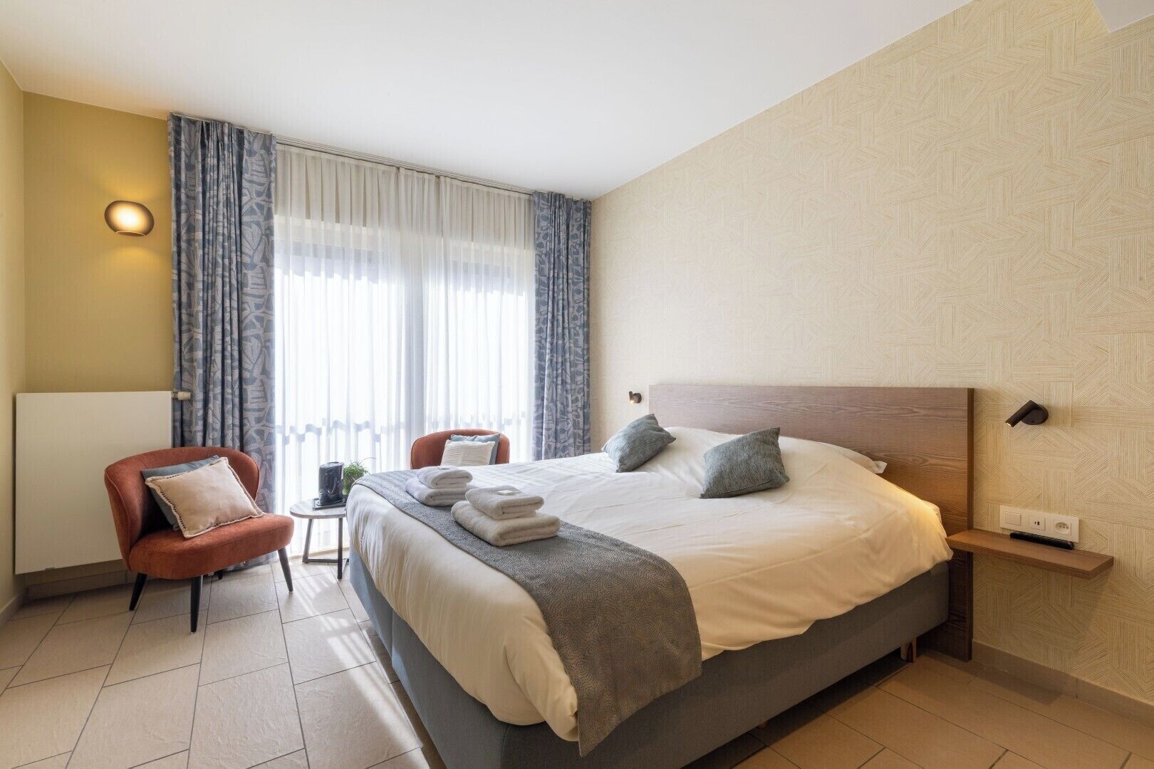 A hotel room with a large bed, two chairs, a bedside table, and a window with curtains.