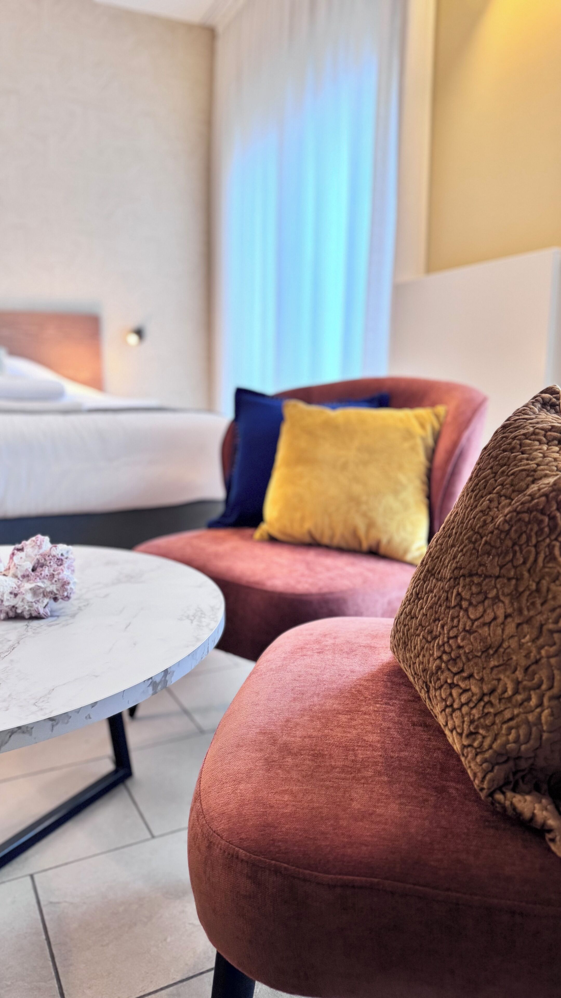Una camera d'albergo moderna con un letto, un tavolino rotondo e un divano rosso con un cuscino giallo.