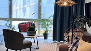 A modern living room with a sofa, armchair, and a floor lamp. There is a large window with a view of buildings and a plant on the windowsill. - Hotel José (Blankenberge)