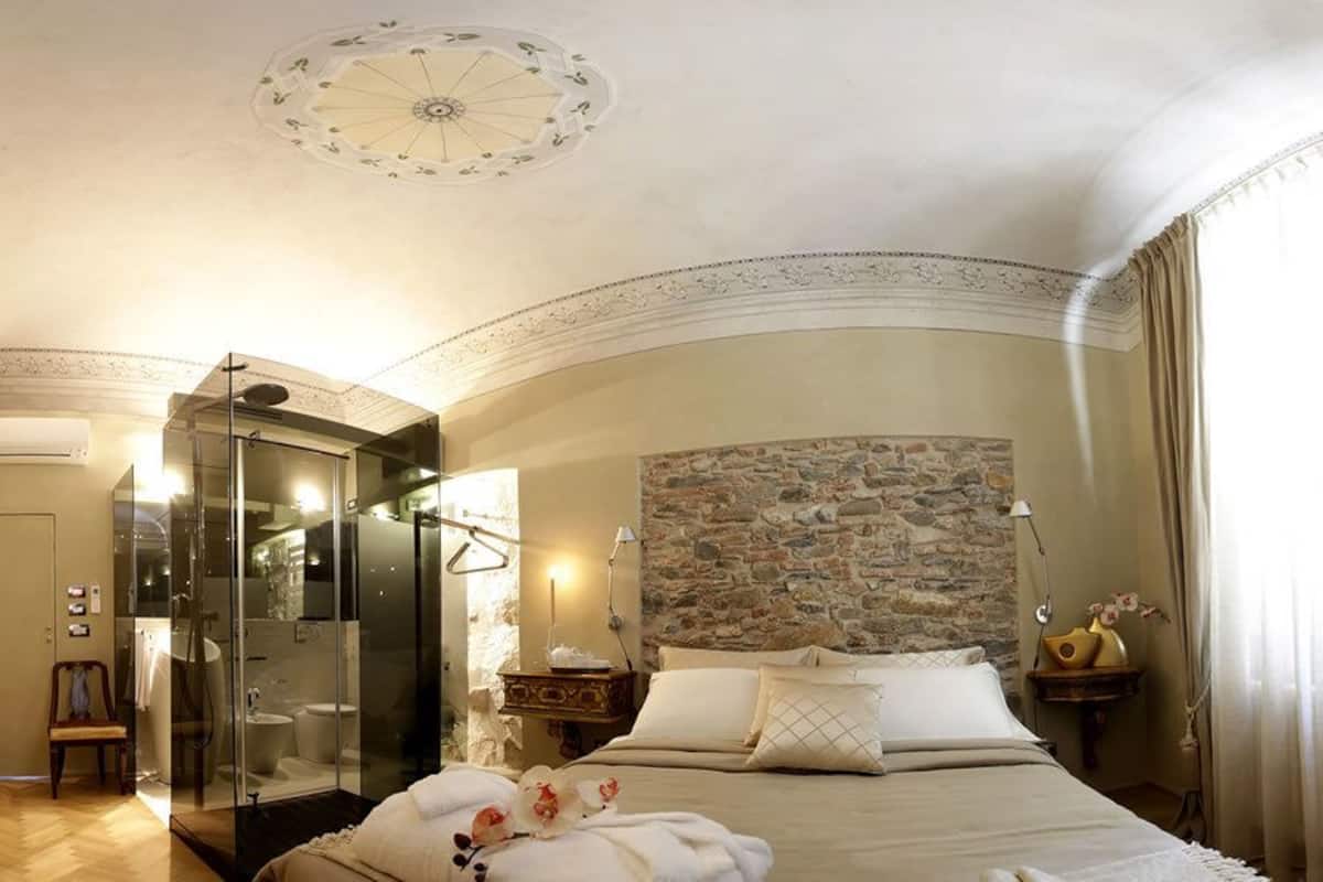A hotel room with a large bed, a stone wall, a glass-enclosed shower, and a chandelier.