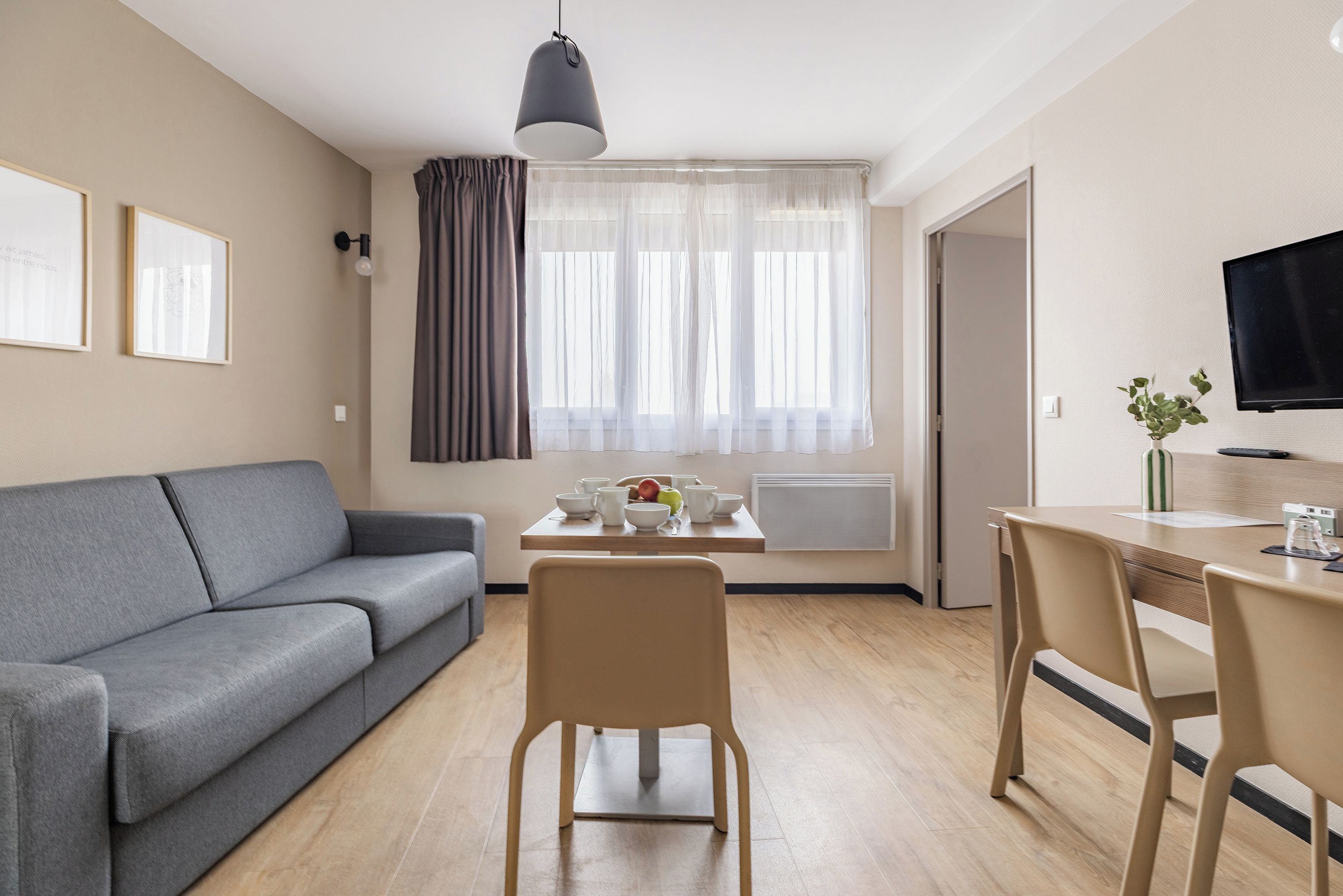 Een moderne woonkamer met een grijze bank, een eettafel met stoelen, een aan de muur bevestigde televisie en een raam met dunne gordijnen.