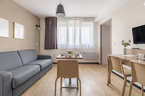 Una sala de estar moderna con un sofá gris, una mesa de comedor con sillas, un televisor montado en la pared y una ventana con cortinas traslúcidas.