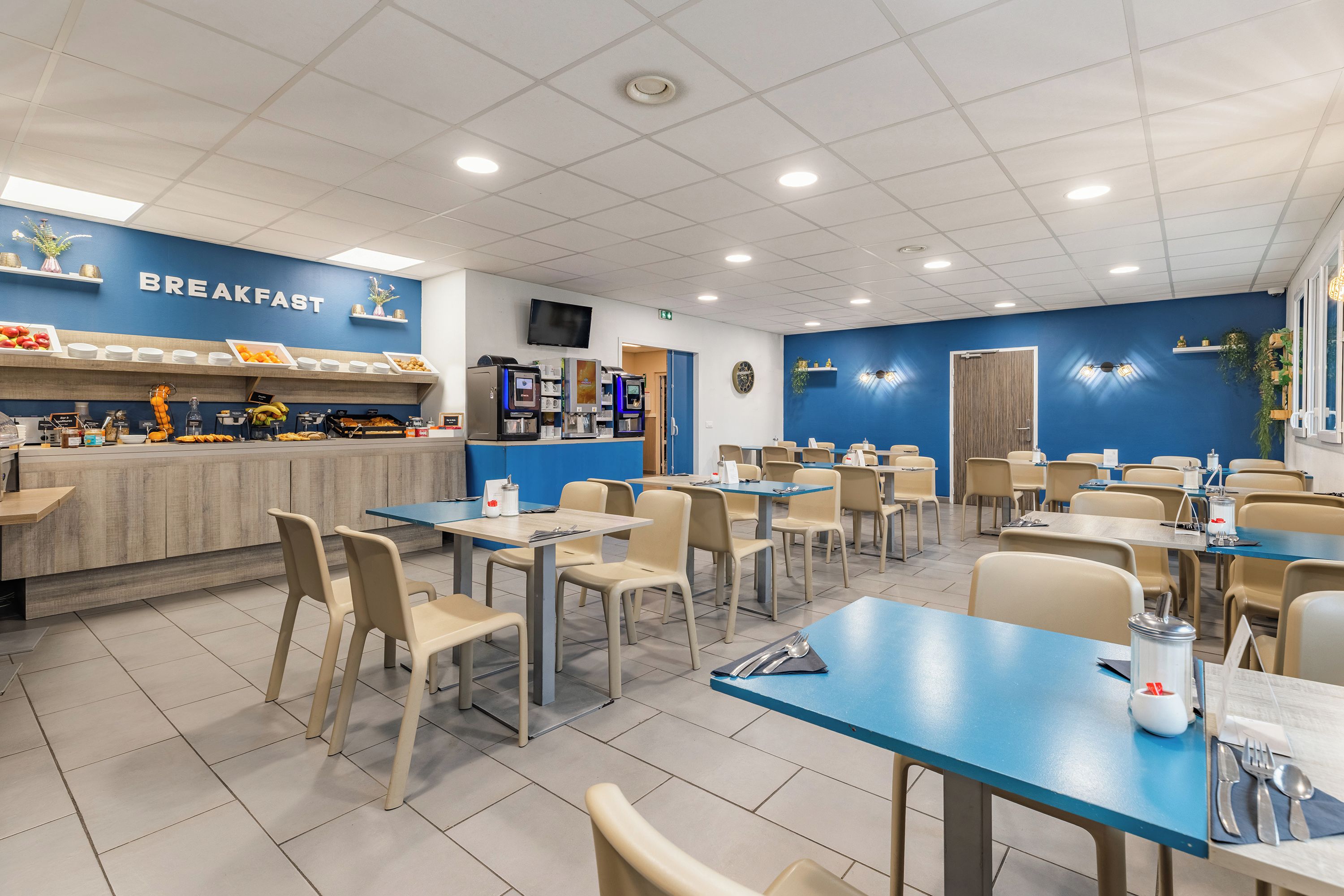 Un espace dédié au petit-déjeuner, avec un mur bleu, un comptoir où sont présentés divers aliments, ainsi que plusieurs tables et chaises.