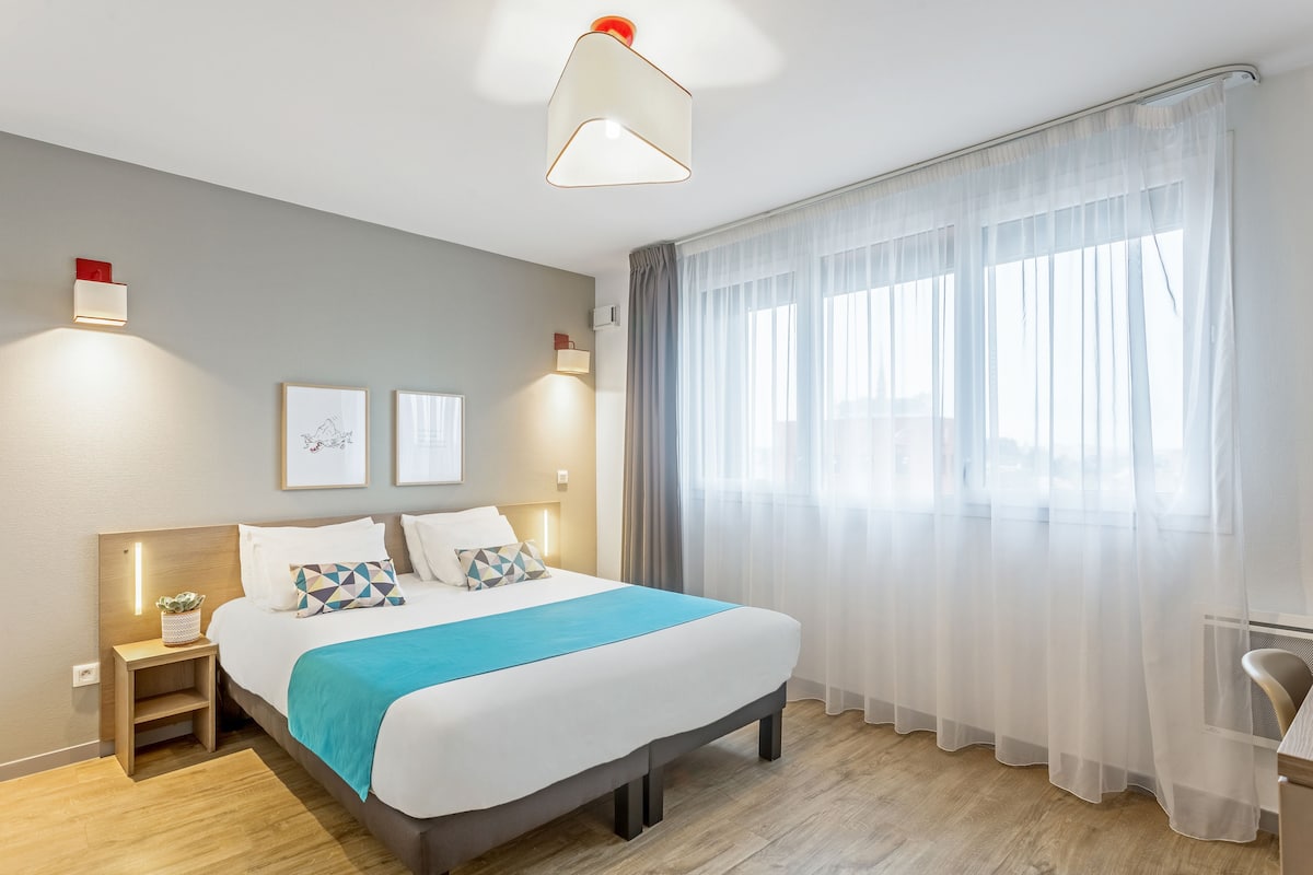 Une chambre d’hôtel moderne dotée d’un grand lit, de tables de chevet, d’un bureau et d’une grande fenêtre avec des rideaux légers.
